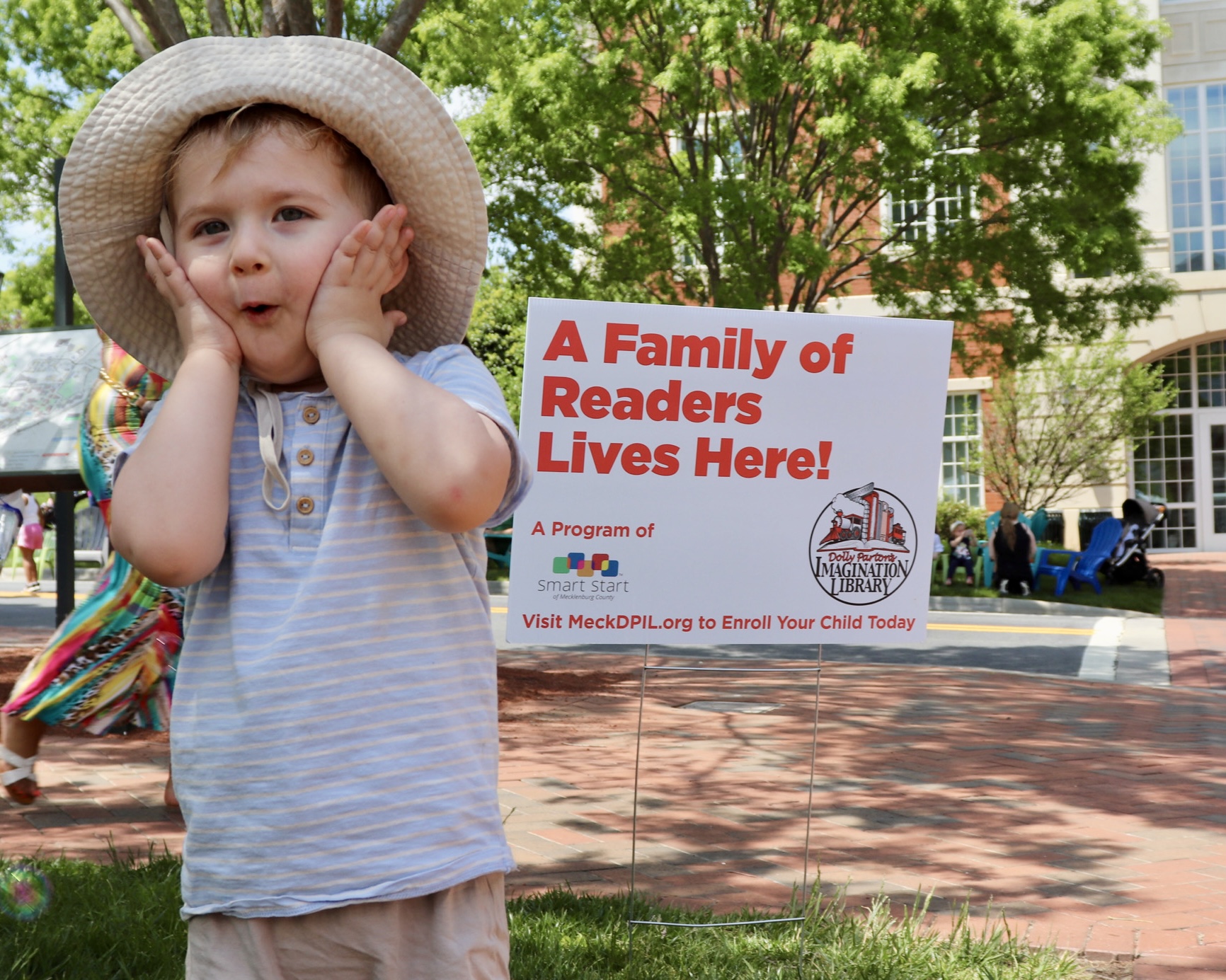 A young child in a sun hat holds both hands to his cheeks beside a yard sign reading "A Family of Readers Lives Here!"