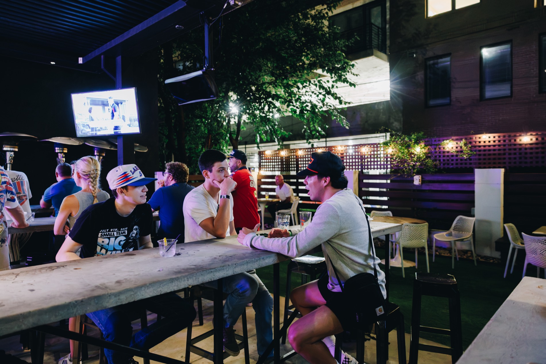 People sit at outdoor picnic tables on a lit patio at night at Hoppin in Charlotte, with a television playing in the background.