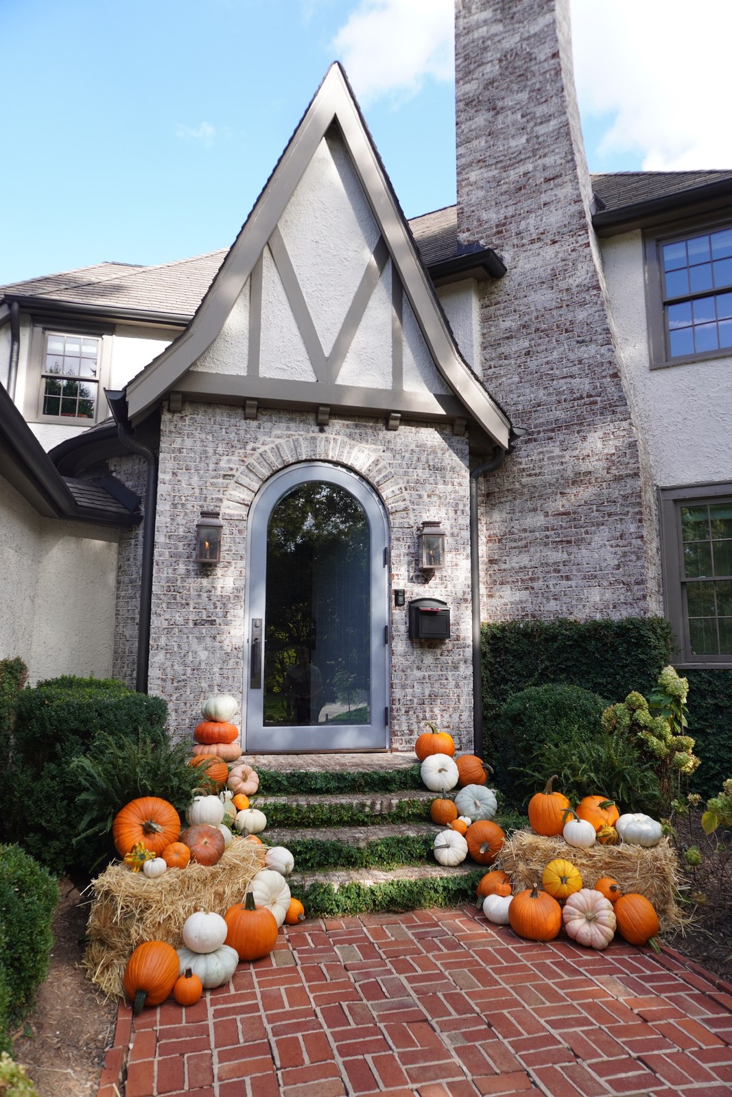 Orange and white pumpkins and hay bales arranged on the front steps of a brick Tudor house