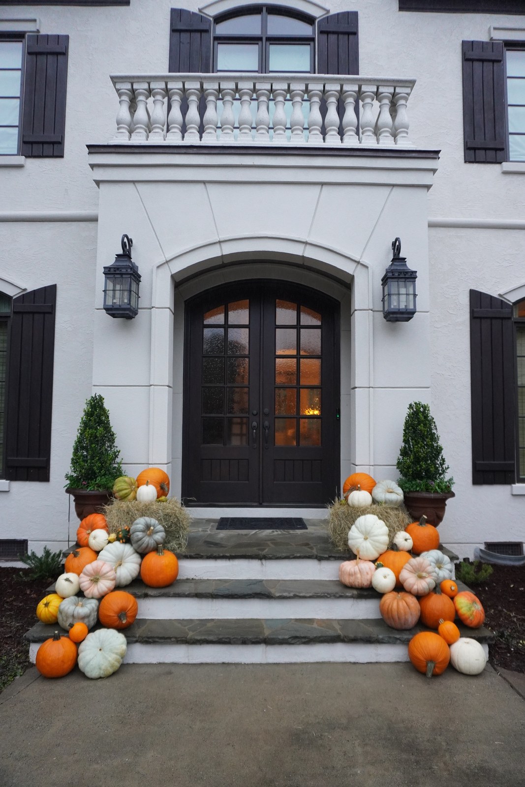Orange, white and green pumpkins with hay bales on the front steps of a white house with dark double doors
