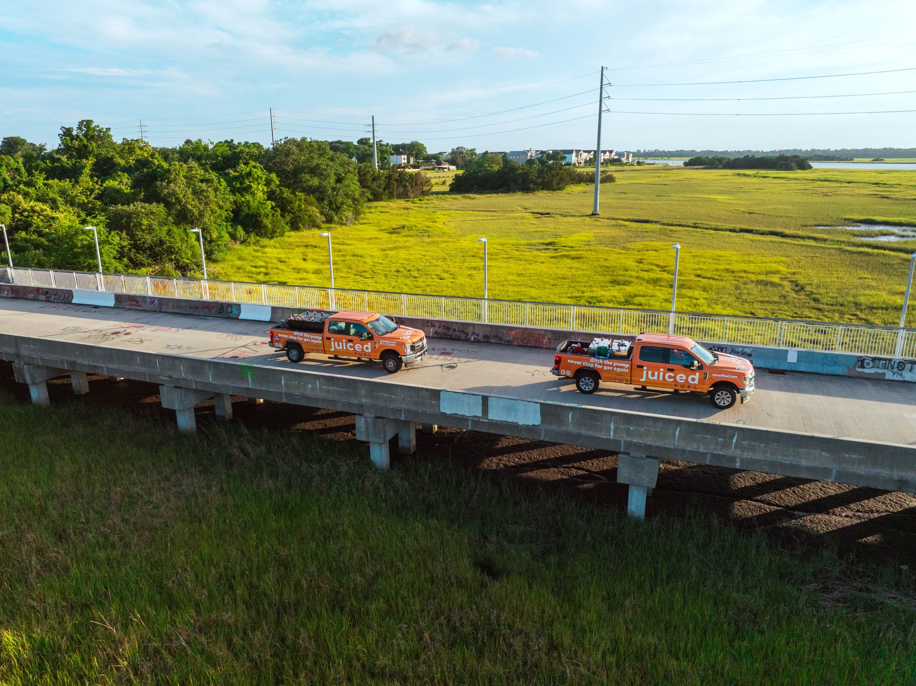 Two orange Juiced Fuel pickup trucks crossing a bridge past a marsh